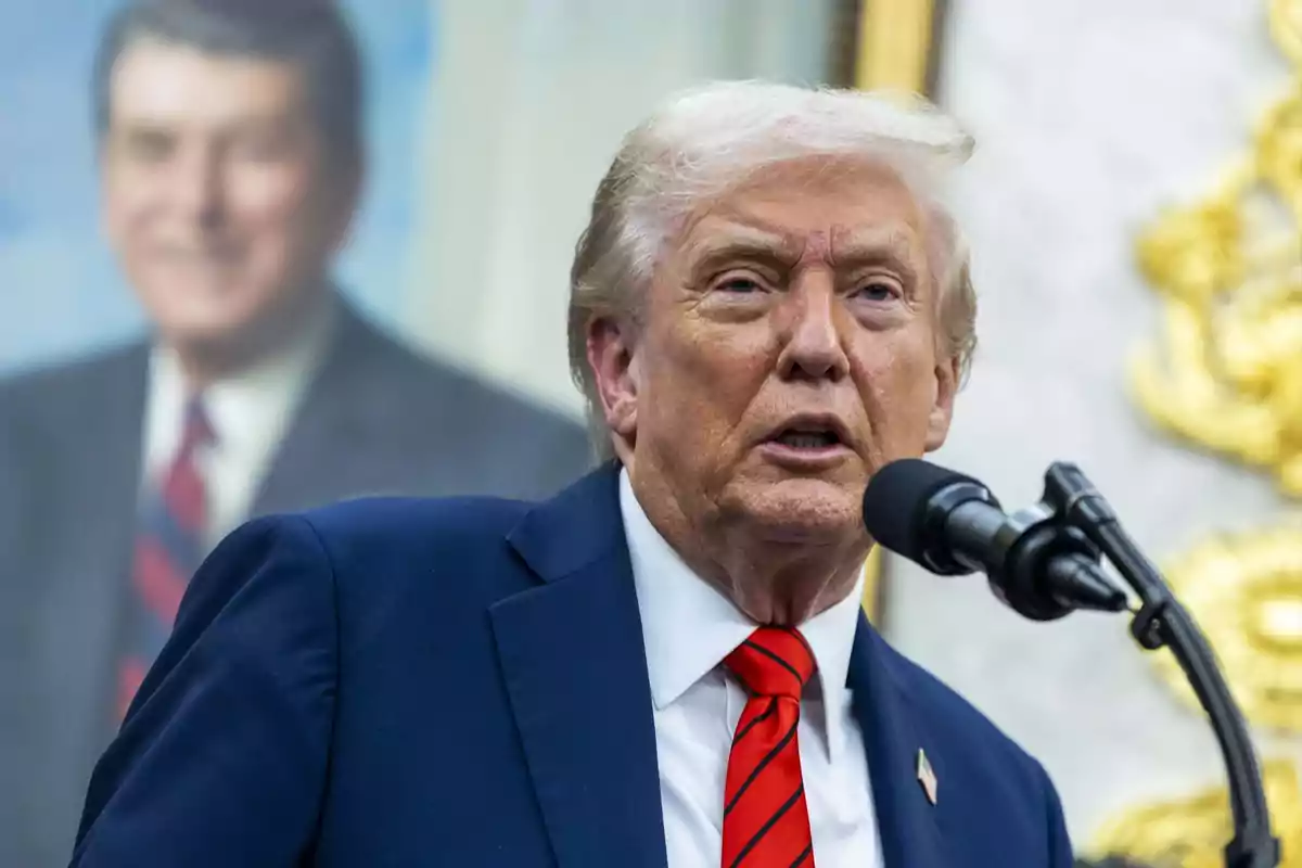 Hombre de cabello rubio con traje azul y corbata roja hablando frente a un micrófono con un retrato borroso al fondo