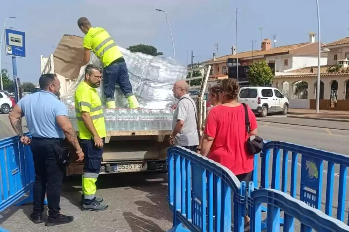 Varias personas recogen botellas de agua de un camión en una calle, rodeados por vallas azules y edificios residenciales al fondo. Varias personas recogen botellas de agua de un camión en una calle, rodeados por vallas azules y edificios residenciales al fondo.