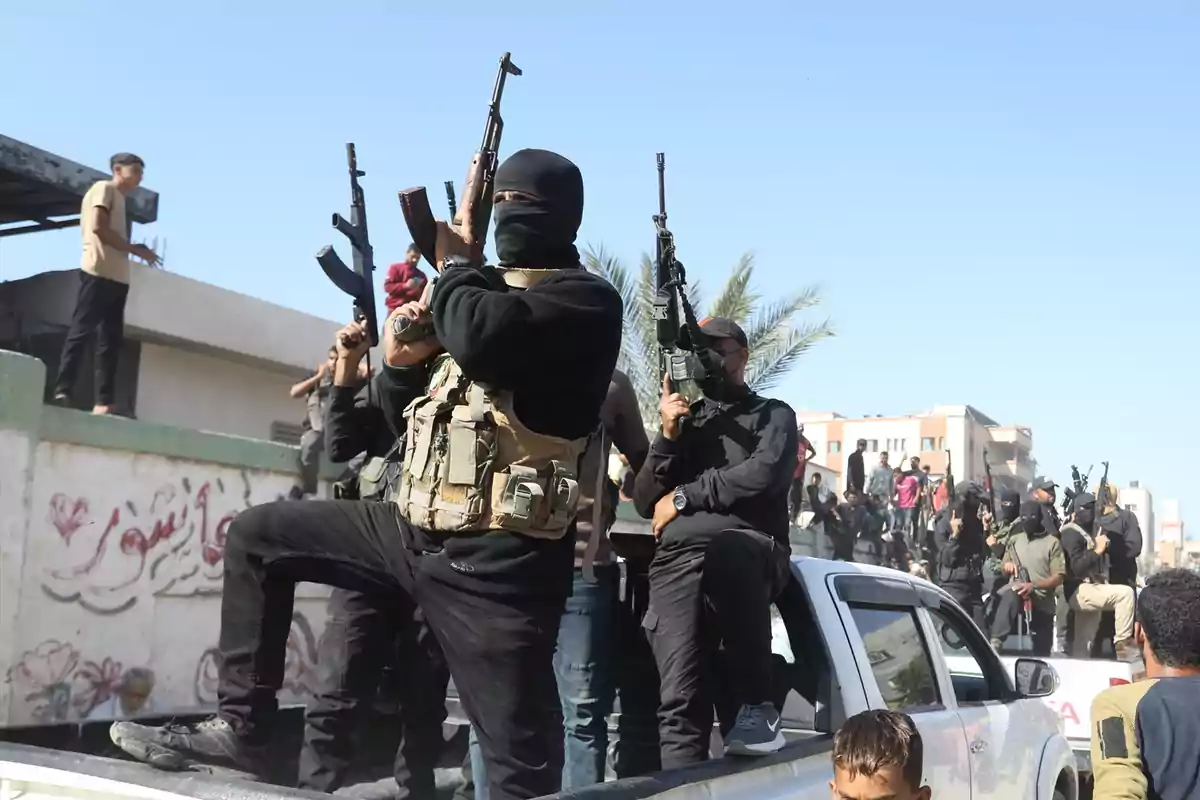 Hombres armados con el rostro cubierto y chalecos tácticos están de pie sobre una camioneta en una calle urbana mientras otras personas observan alrededor