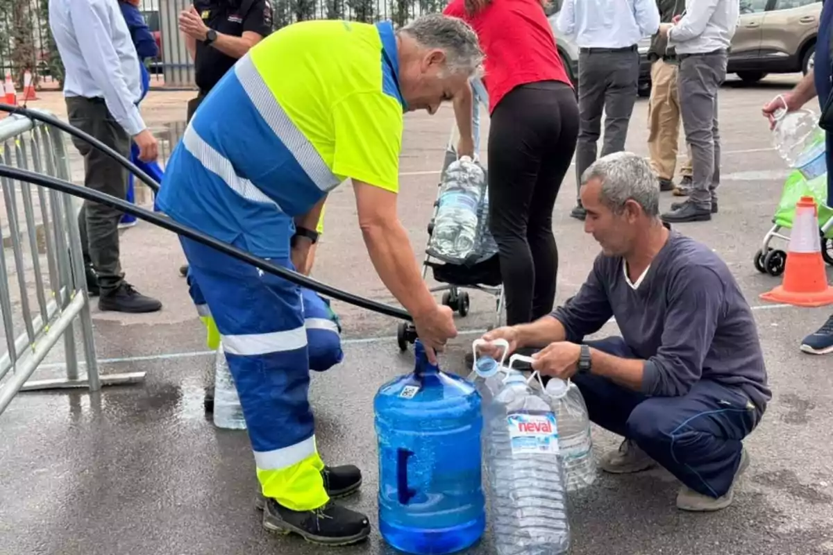 Un trabajador con uniforme de alta visibilidad llena garrafones de agua mientras un hombre sostiene botellas vacías en una zona de reparto de agua, rodeados de más personas y vehículos en el fondo.