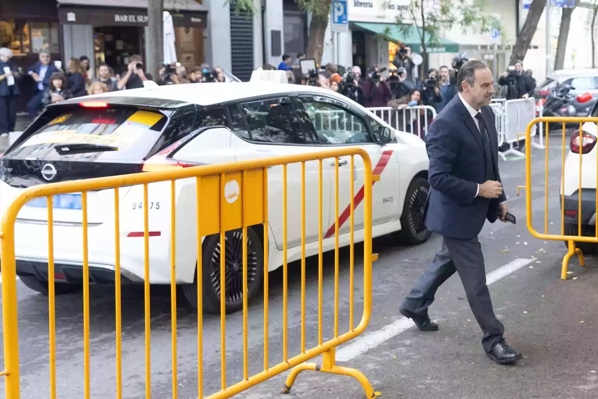 Un hombre con traje camina por la calle junto a unas vallas amarillas y un taxi blanco mientras varias personas y fotógrafos observan al fondo Un hombre con traje camina por la calle junto a unas vallas amarillas y un taxi blanco mientras varias personas y fotógrafos observan al fondo