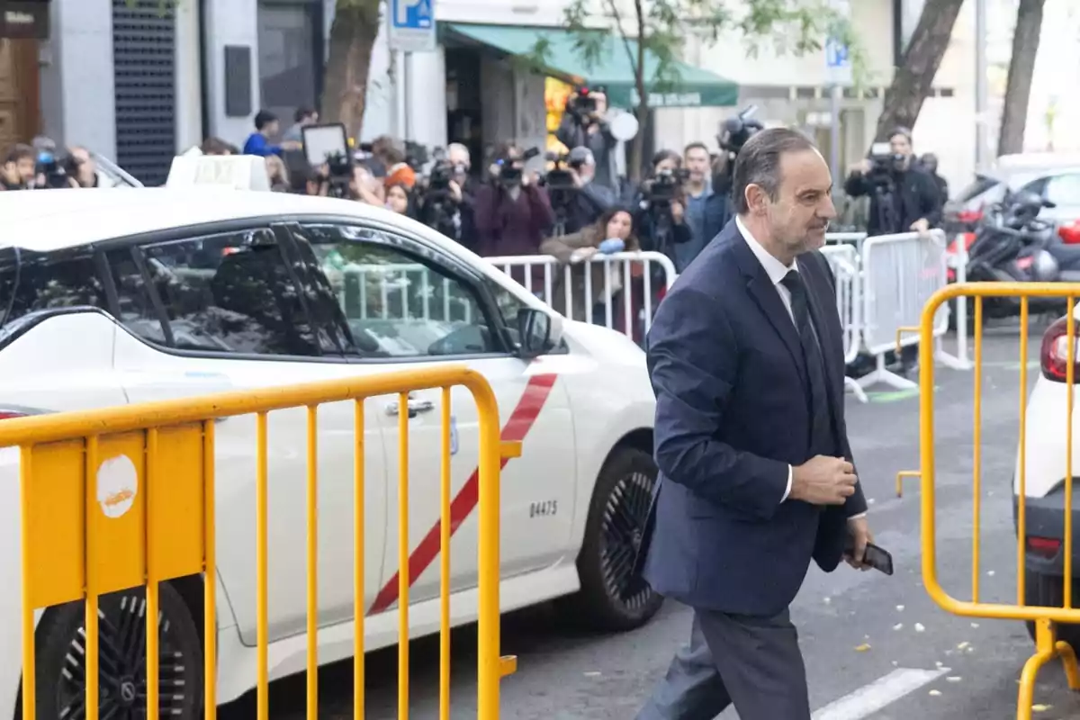 Hombre con traje caminando junto a una valla amarilla frente a un taxi blanco mientras un grupo de periodistas y fotógrafos lo observa al fondo