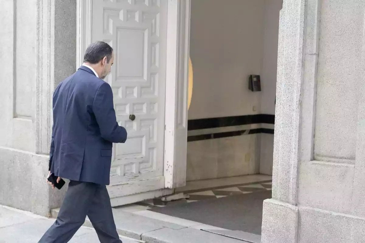Hombre de traje azul entrando a un edificio por una puerta blanca mientras sostiene un teléfono móvil en la mano derecha Hombre de traje azul entrando a un edificio por una puerta blanca mientras sostiene un teléfono móvil en la mano derecha