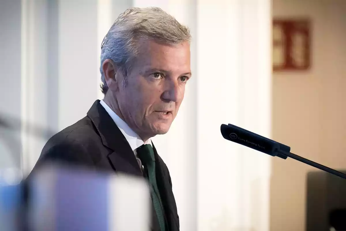 Hombre de cabello canoso con traje oscuro hablando frente a un micrófono en un entorno profesional