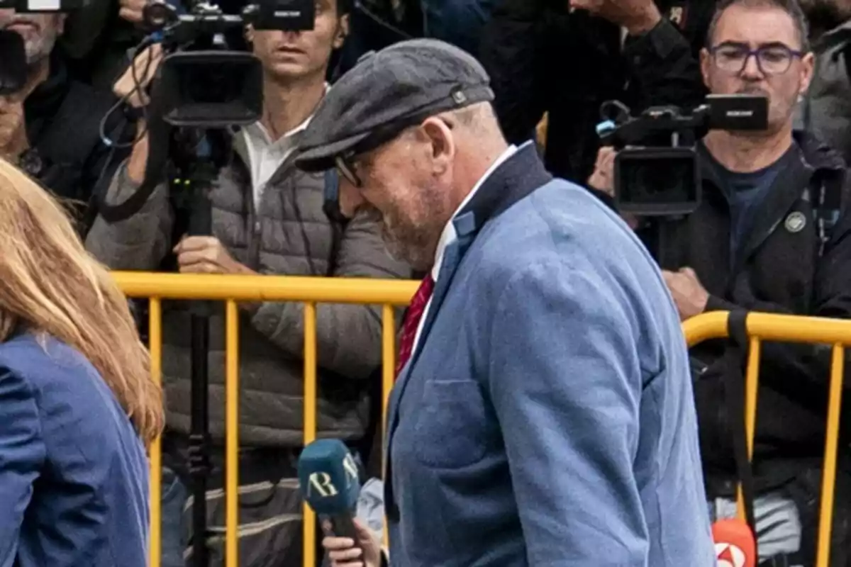 Hombre con gorra y gafas camina frente a una valla amarilla mientras varios camarógrafos lo graban