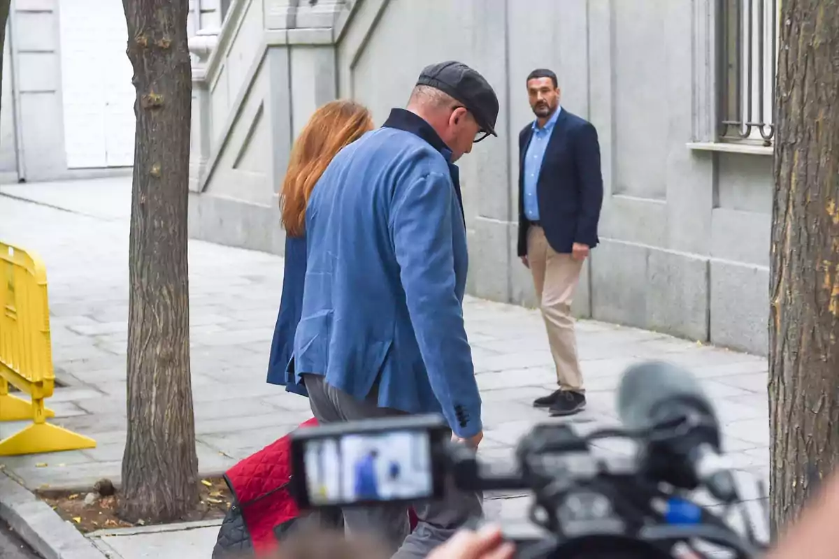 Tres personas en una acera junto a un edificio gris, una cámara de televisión en primer plano y barreras amarillas al fondo Tres personas en una acera junto a un edificio gris, una cámara de televisión en primer plano y barreras amarillas al fondo