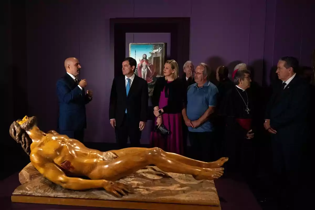 Un grupo de personas elegantemente vestidas observa una escultura de Cristo y una pintura religiosa en una sala de museo con paredes moradas Un grupo de personas elegantemente vestidas observa una escultura de Cristo y una pintura religiosa en una sala de museo con paredes moradas