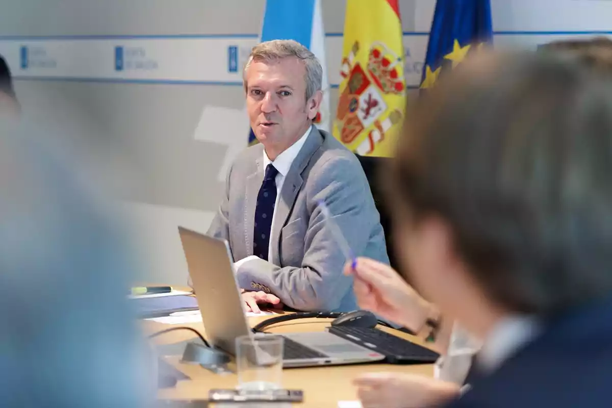 Un hombre de cabello canoso con traje gris y corbata azul está sentado en una mesa de reuniones con portátiles y documentos, al fondo se ven banderas de España, Galicia y la Unión Europea. Un hombre de cabello canoso con traje gris y corbata azul está sentado en una mesa de reuniones con portátiles y documentos, al fondo se ven banderas de España, Galicia y la Unión Europea.