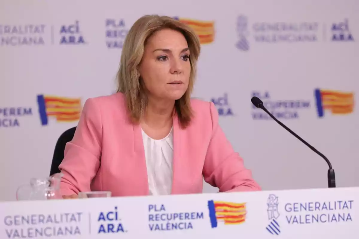 Mujer con chaqueta rosa sentada en una mesa durante una rueda de prensa con fondo de logotipos de la Generalitat Valenciana
