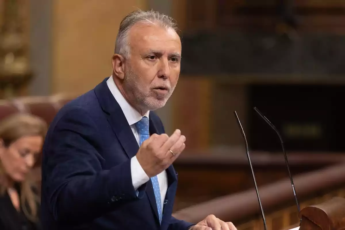 Hombre de cabello canoso y barba hablando en un podio con micrófonos en un entorno parlamentario