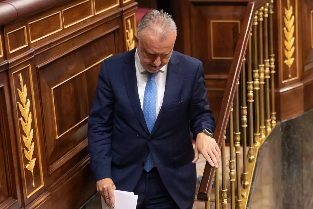 Hombre de cabello canoso con traje azul y corbata celeste bajando unas escaleras de madera decoradas con detalles dorados en un entorno institucional