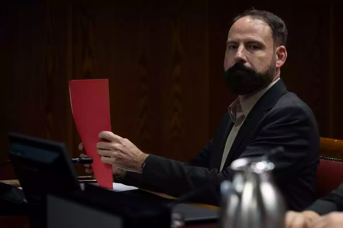 Hombre de barba y traje oscuro sentado en una mesa de madera sosteniendo una carpeta roja mientras mira hacia el frente