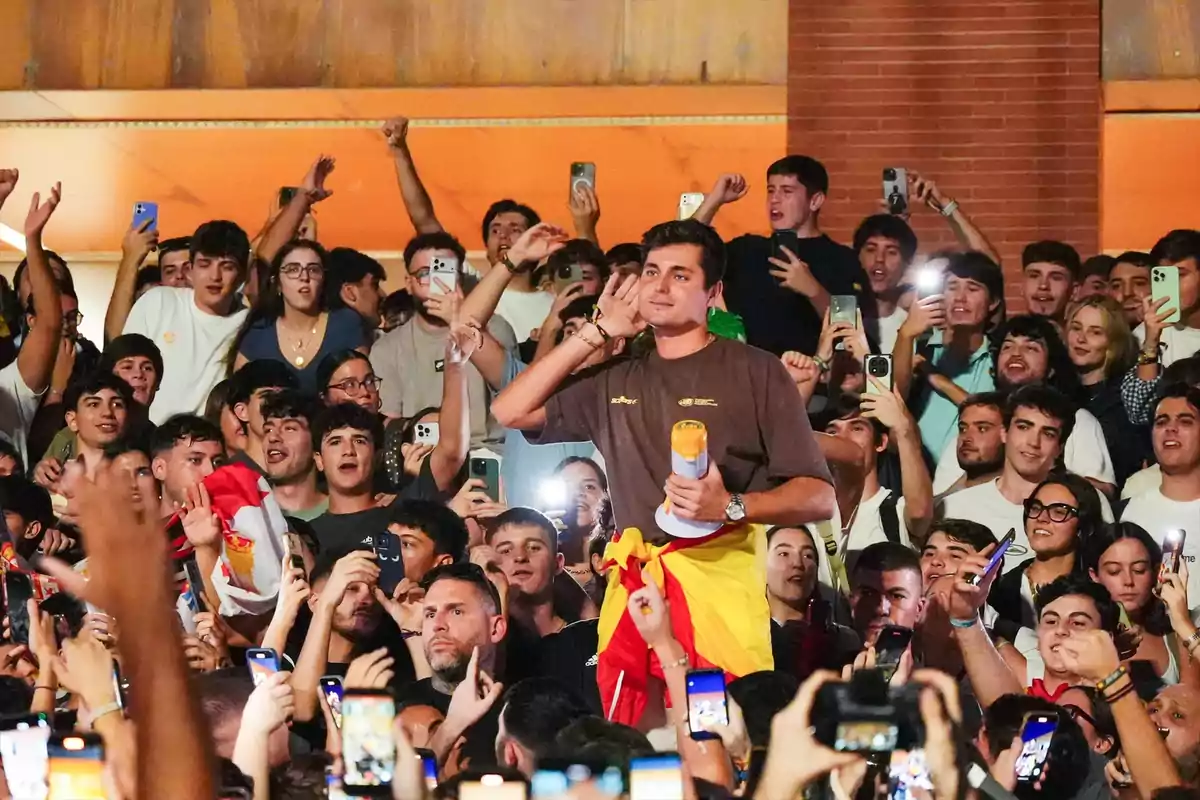 Un joven envuelto en una bandera es levantado por una multitud de personas que lo animan y graban con sus teléfonos móviles durante un evento festivo