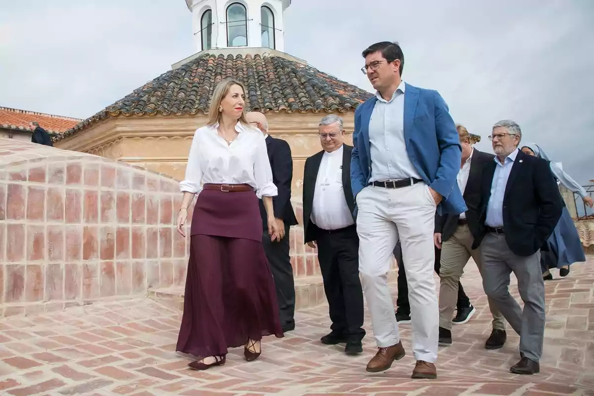 Un grupo de personas camina sobre una superficie de ladrillo junto a un edificio con techo de tejas y una cúpula blanca al fondo