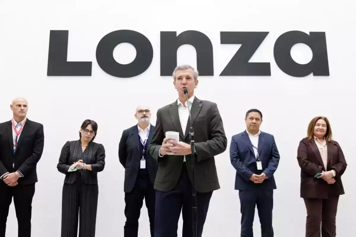 Un grupo de seis personas vestidas de manera formal está de pie frente a una pared blanca con el logotipo de Lonza mientras una de ellas habla al micrófono