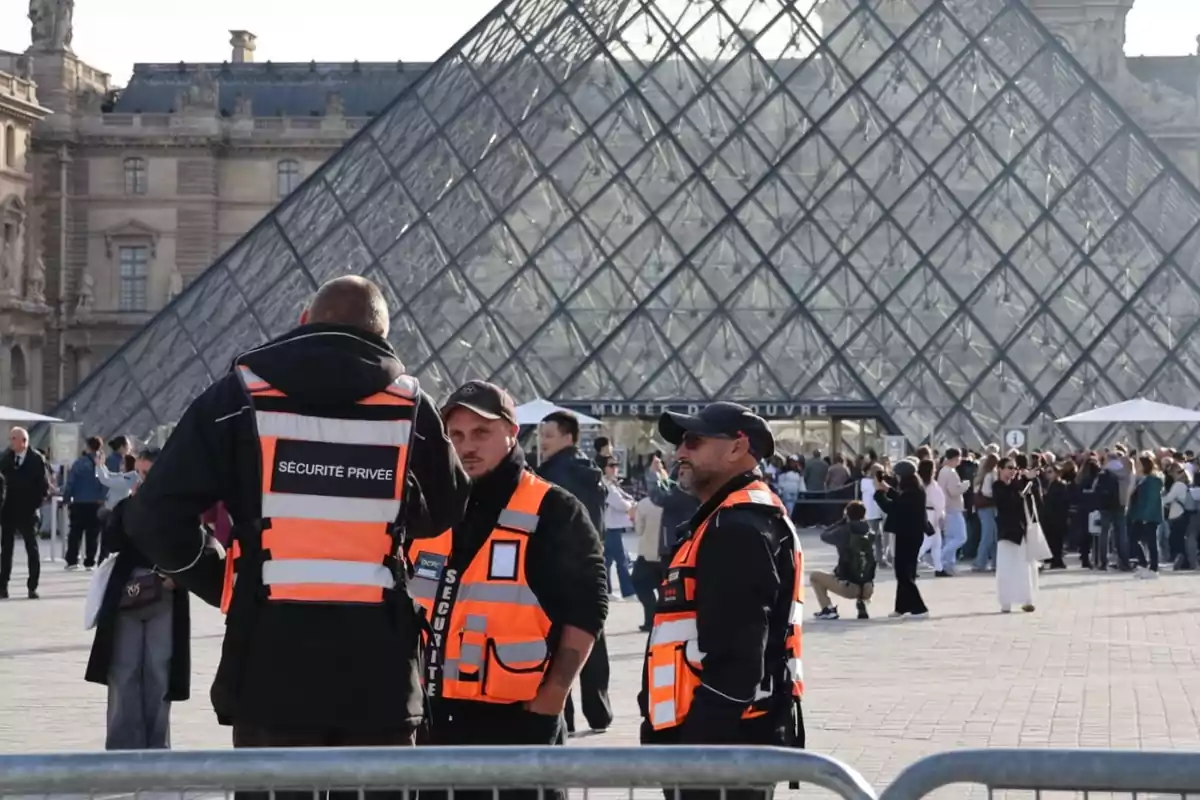 Tres guardias de seguridad con chalecos naranjas están de pie frente a la pirámide de cristal del Museo del Louvre mientras varias personas hacen fila en el exterior Tres guardias de seguridad con chalecos naranjas están de pie frente a la pirámide de cristal del Museo del Louvre mientras varias personas hacen fila en el exterior