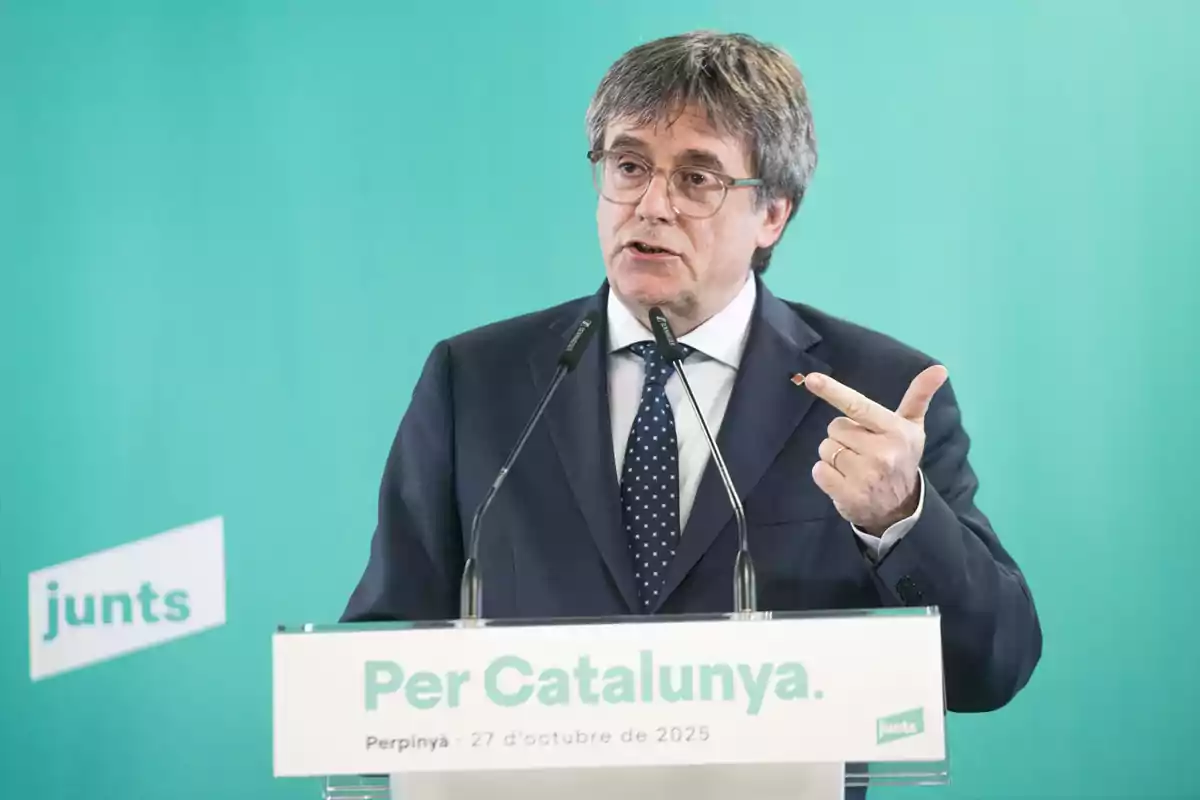 Hombre con gafas y traje hablando en un atril con el texto Per Catalunya sobre un fondo turquesa