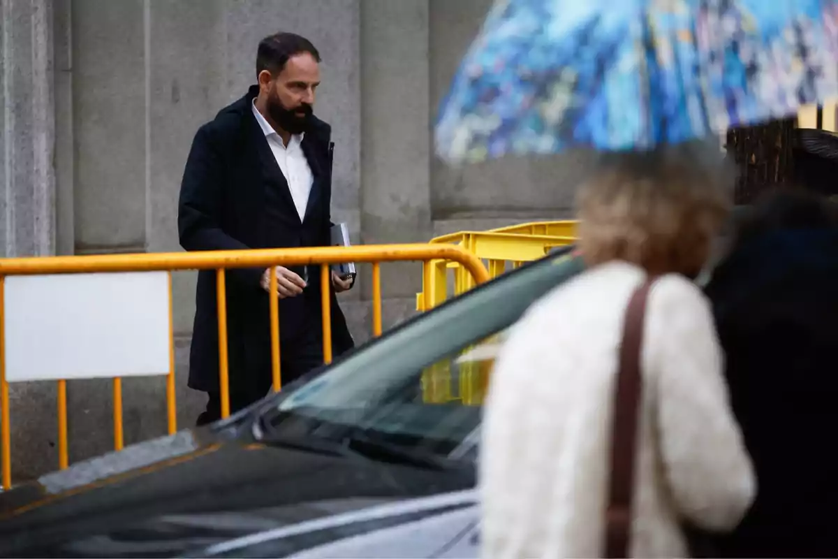 Hombre con barba y abrigo oscuro camina junto a una valla amarilla mientras una persona con paraguas colorido pasa por delante Hombre con barba y abrigo oscuro camina junto a una valla amarilla mientras una persona con paraguas colorido pasa por delante
