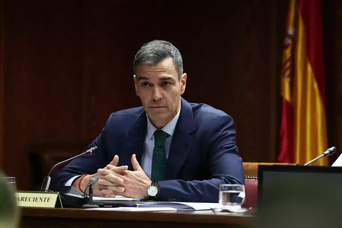 Hombre con traje azul y corbata verde sentado en una mesa de reuniones con micrófono y bandera de España al fondo