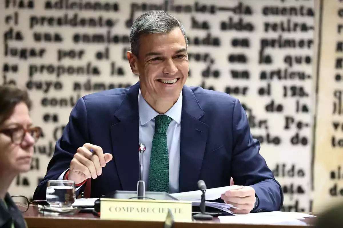 Hombre con traje azul y corbata verde sonríe mientras sostiene unos papeles en una mesa de conferencia con micrófonos y un cartel que dice compareciente al frente Hombre con traje azul y corbata verde sonríe mientras sostiene unos papeles en una mesa de conferencia con micrófonos y un cartel que dice compareciente al frente