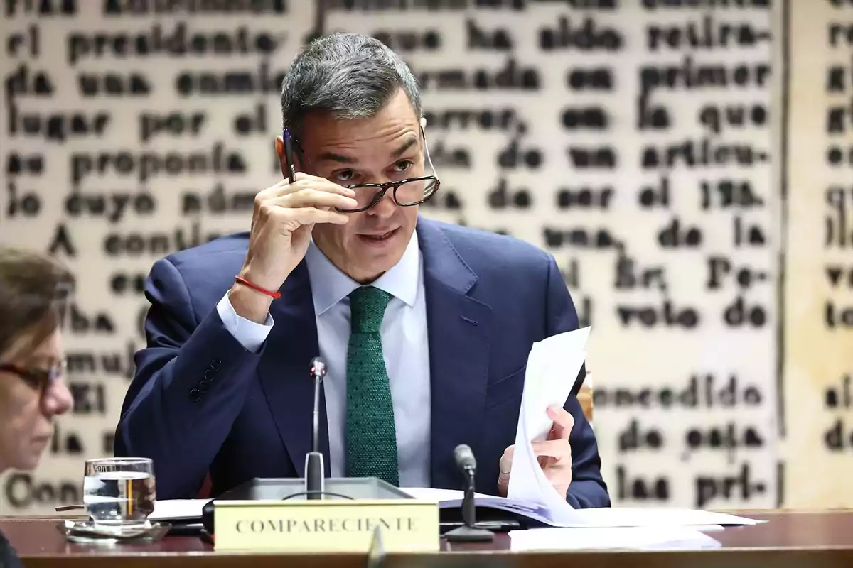 Hombre de traje azul y corbata verde ajustándose las gafas mientras sostiene unos papeles en una comparecencia oficial con un cartel que dice compareciente y fondo de letras desenfocadas