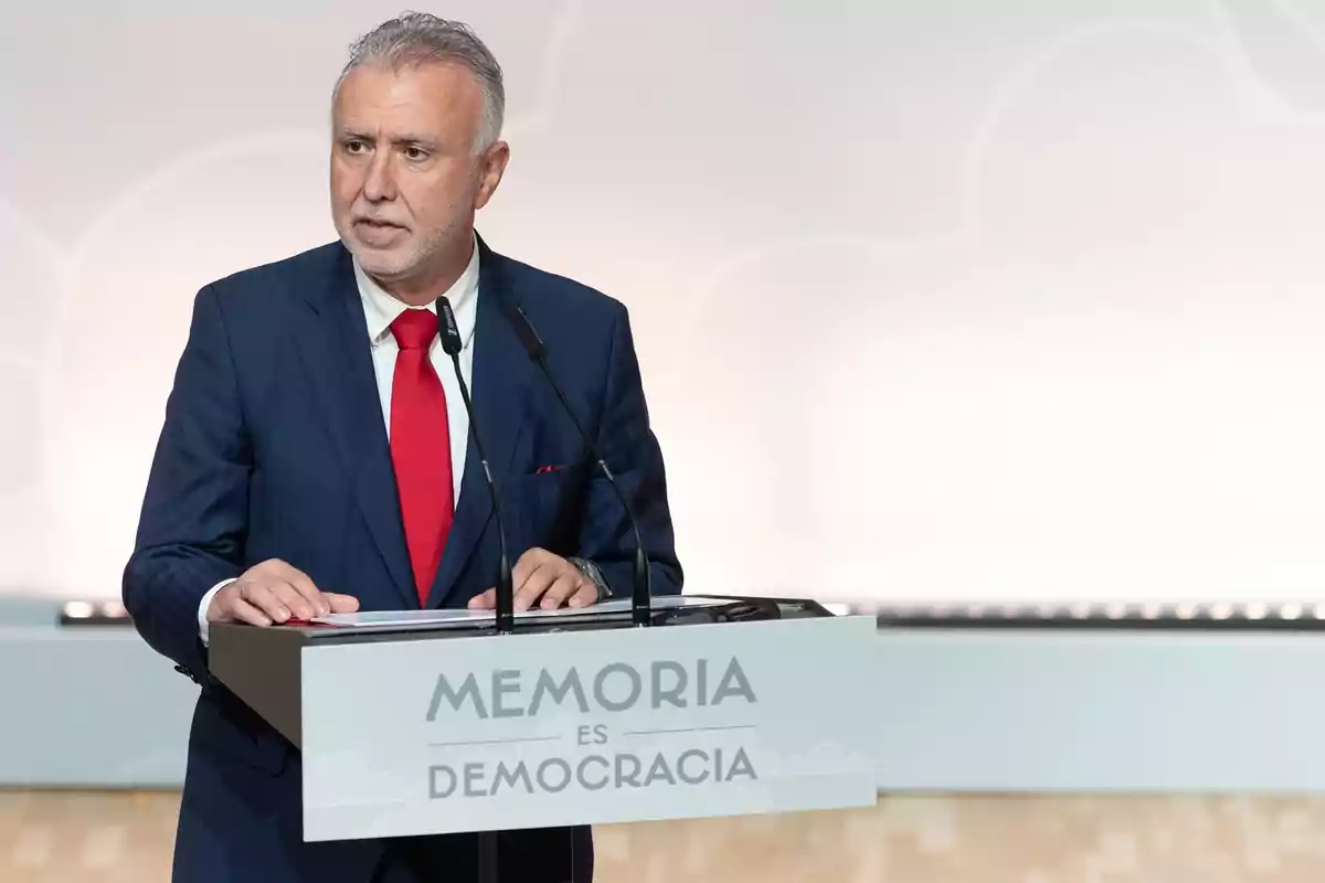 Hombre de cabello canoso con traje azul y corbata roja hablando en un atril que tiene el texto Memoria es democracia Hombre de cabello canoso con traje azul y corbata roja hablando en un atril que tiene el texto Memoria es democracia