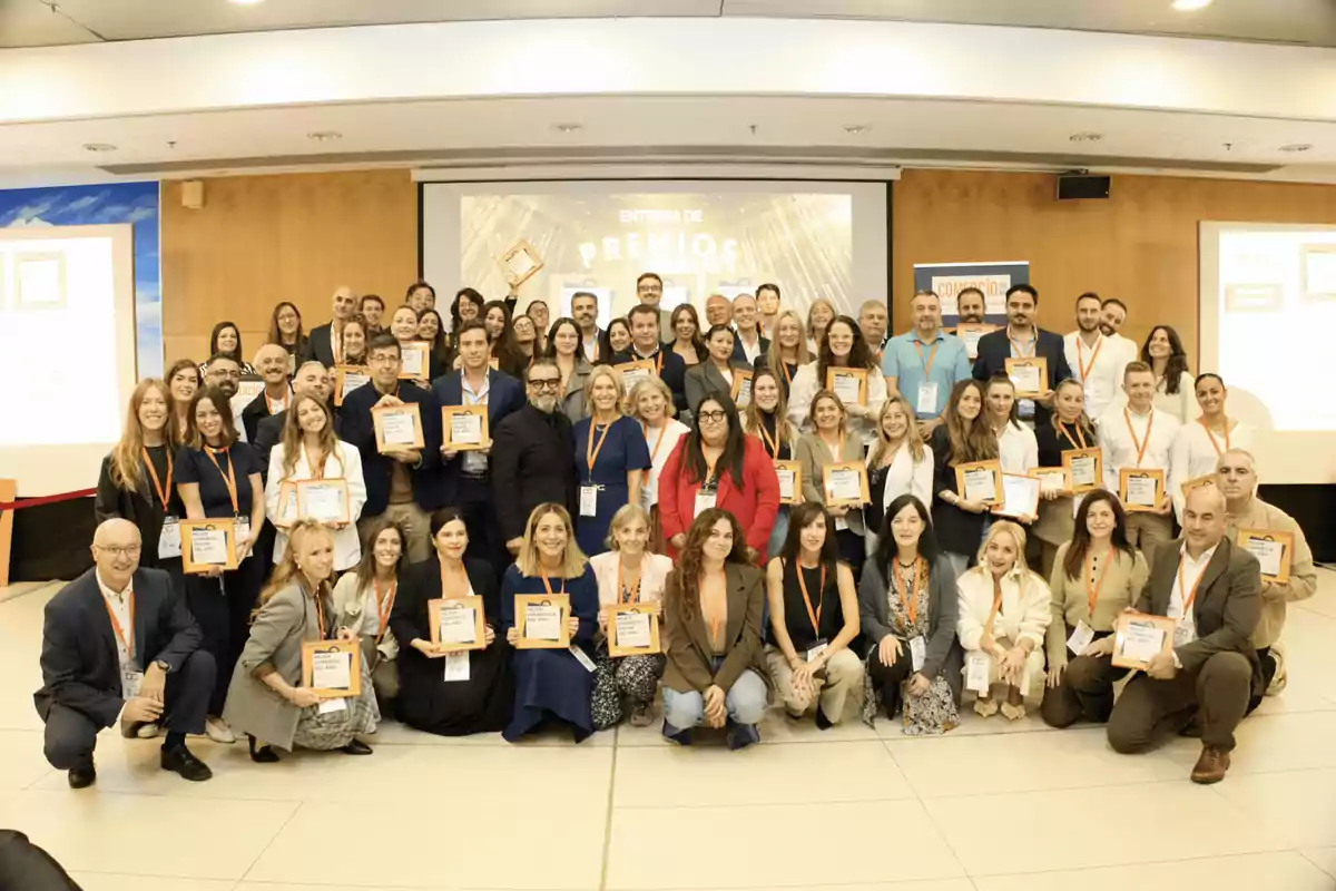 Grupo numeroso de personas posando juntas en un salón de eventos, muchas de ellas sosteniendo placas de reconocimiento y usando gafetes naranjas, con una pantalla al fondo que muestra la palabra premios.
