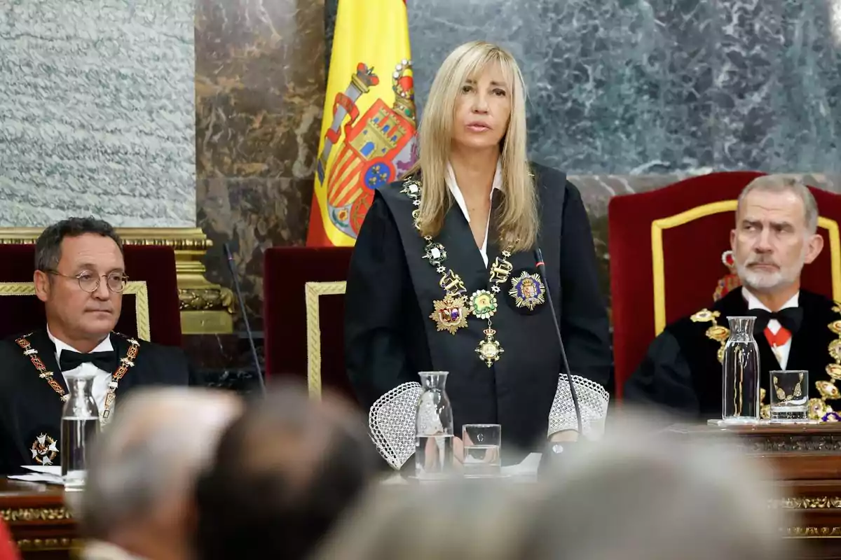 Una mujer con toga y medallas habla de pie en un acto oficial junto a dos hombres sentados, con una bandera de España al fondo.
