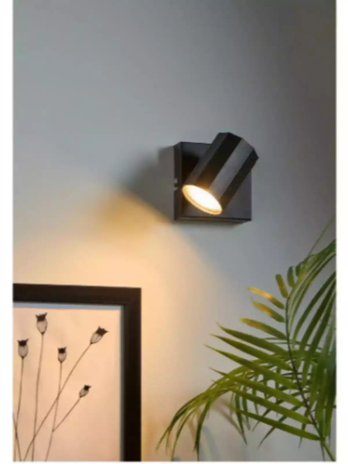 Lámpara de pared negra encendida iluminando una pared junto a una planta y un cuadro decorativo