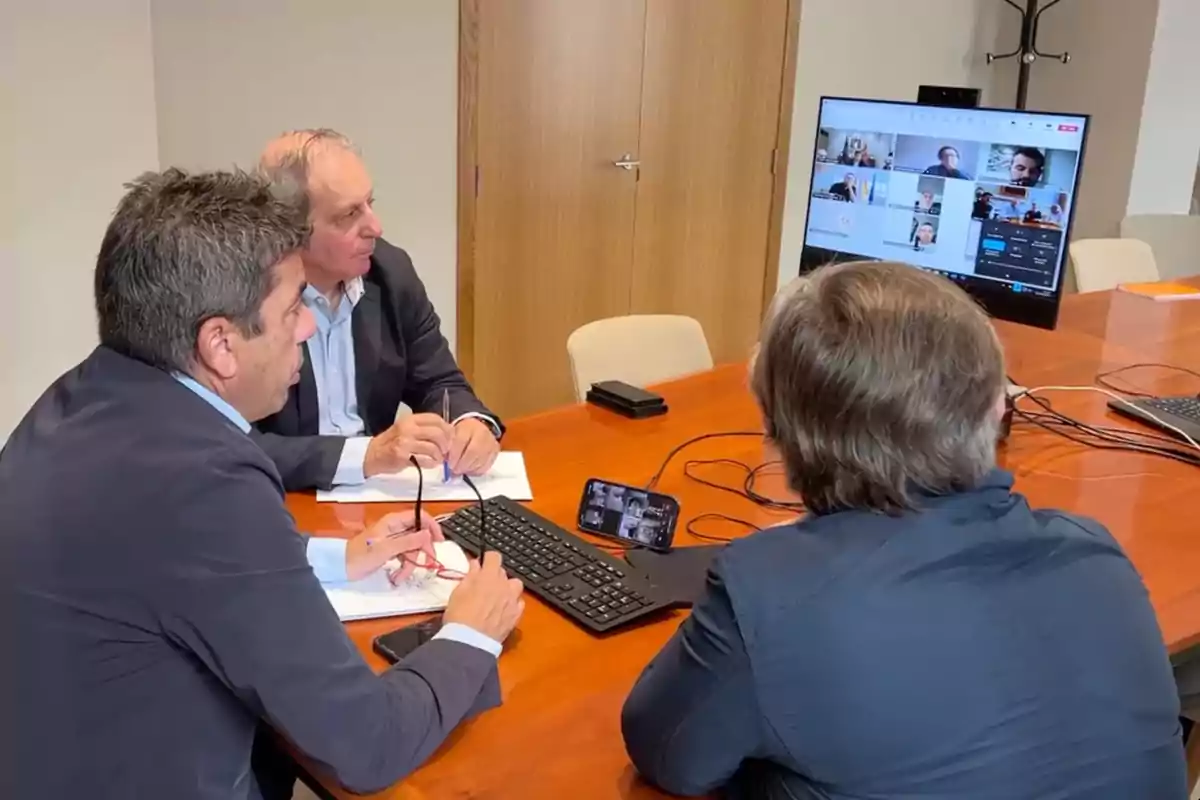 Tres hombres sentados alrededor de una mesa de madera participan en una videollamada grupal usando una computadora y un teléfono móvil Tres hombres sentados alrededor de una mesa de madera participan en una videollamada grupal usando una computadora y un teléfono móvil