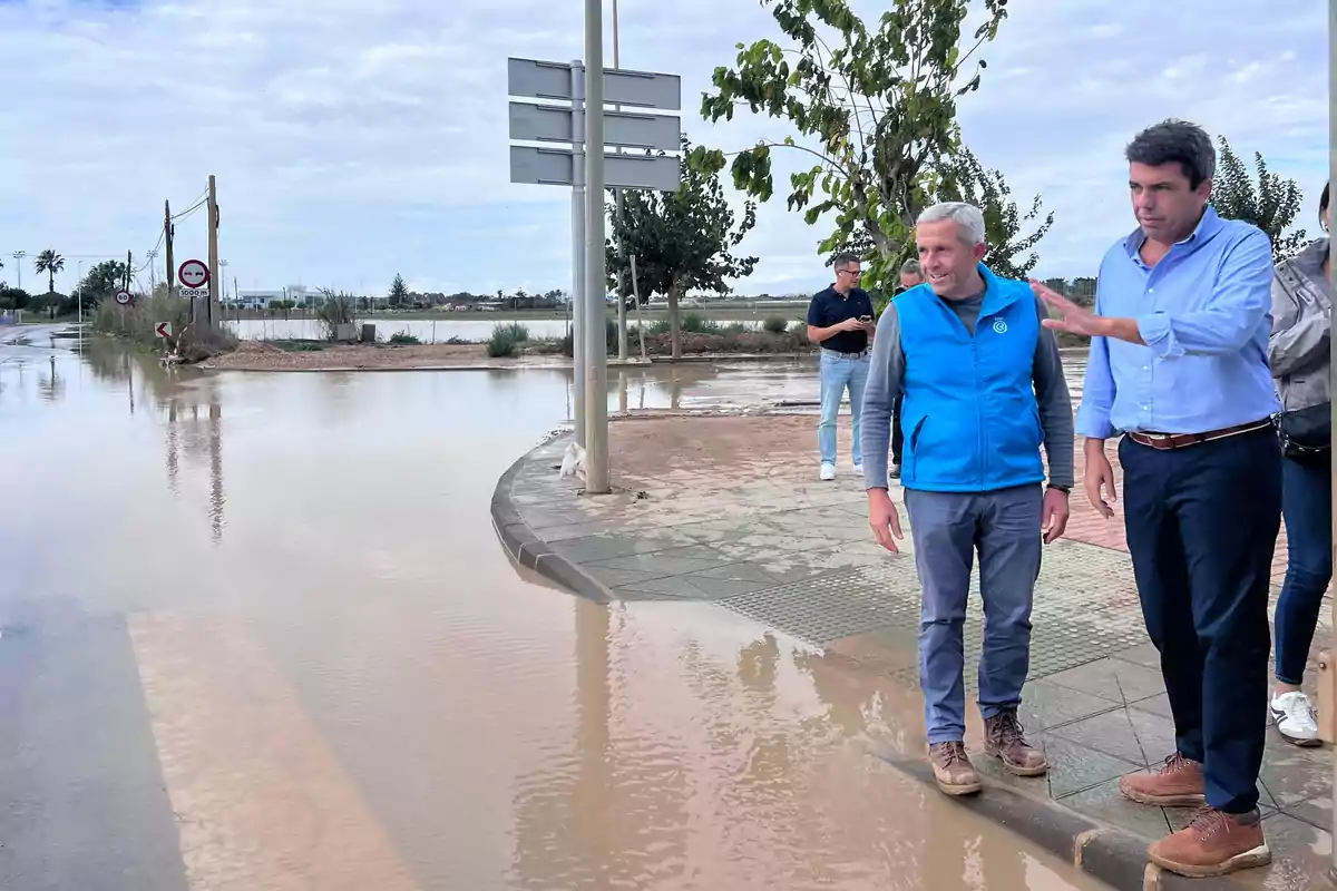 Varias personas observan una calle inundada mientras permanecen en la acera junto a un charco grande de agua turbia Varias personas observan una calle inundada mientras permanecen en la acera junto a un charco grande de agua turbia