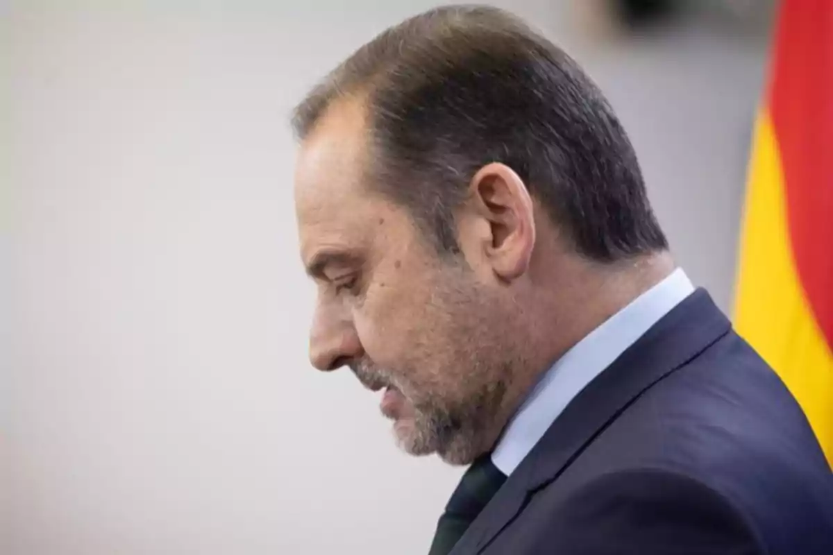 Hombre de perfil con expresión seria vistiendo traje oscuro y corbata frente a una bandera desenfocada en el fondo Hombre de perfil con expresión seria vistiendo traje oscuro y corbata frente a una bandera desenfocada en el fondo