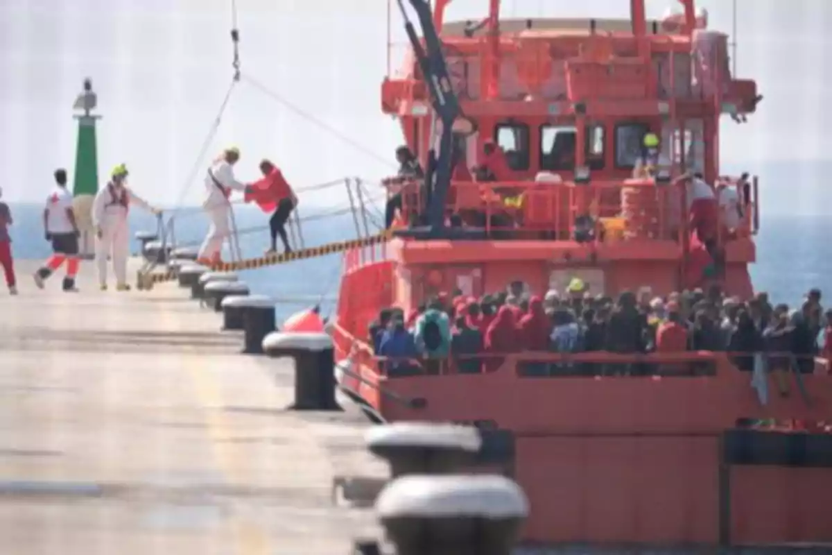 Un barco de rescate naranja llega al muelle con muchas personas a bordo mientras algunas personas con chalecos y personal de ayuda asisten en el desembarco Un barco de rescate naranja llega al muelle con muchas personas a bordo mientras algunas personas con chalecos y personal de ayuda asisten en el desembarco