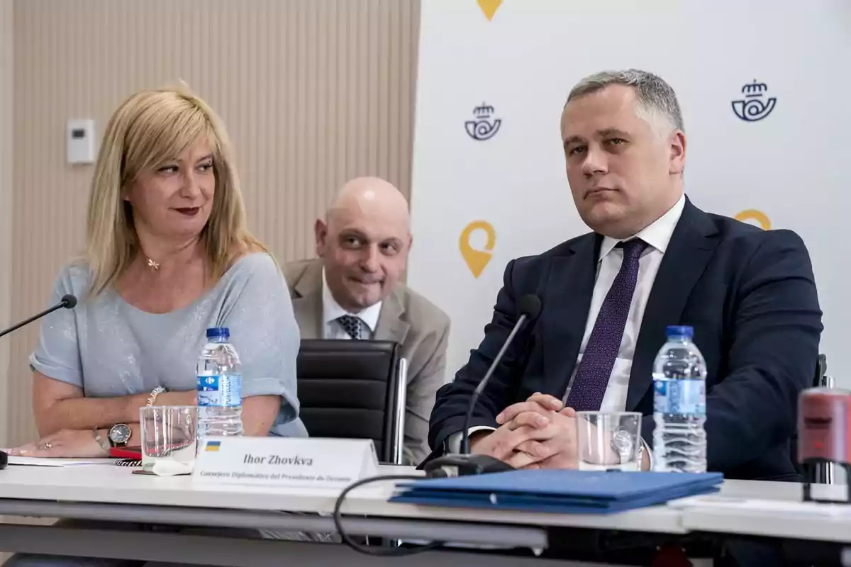 Tres personas sentadas en una mesa de conferencia con micrófonos y botellas de agua, al fondo un cartel con logotipos institucionales. Tres personas sentadas en una mesa de conferencia con micrófonos y botellas de agua, al fondo un cartel con logotipos institucionales.