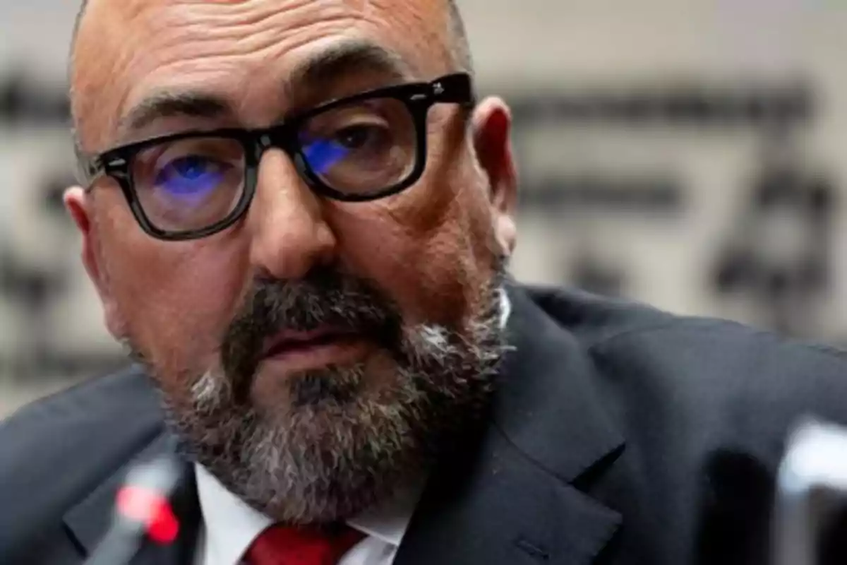 Hombre de mediana edad con barba y gafas, vestido con traje oscuro y corbata roja, mirando hacia el frente en un entorno profesional Hombre de mediana edad con barba y gafas, vestido con traje oscuro y corbata roja, mirando hacia el frente en un entorno profesional