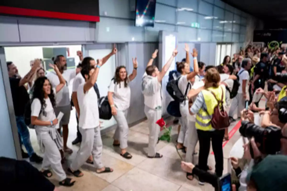Un grupo de personas vestidas de blanco es recibido con entusiasmo por una multitud en un aeropuerto mientras algunos levantan los brazos y otros toman fotos