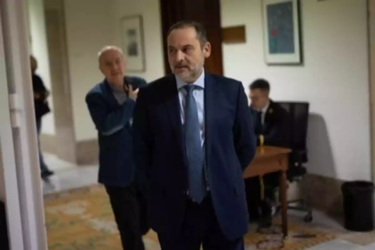 Hombre de traje azul caminando por un pasillo con otras personas al fondo en un ambiente de oficina o institucional Hombre de traje azul caminando por un pasillo con otras personas al fondo en un ambiente de oficina o institucional