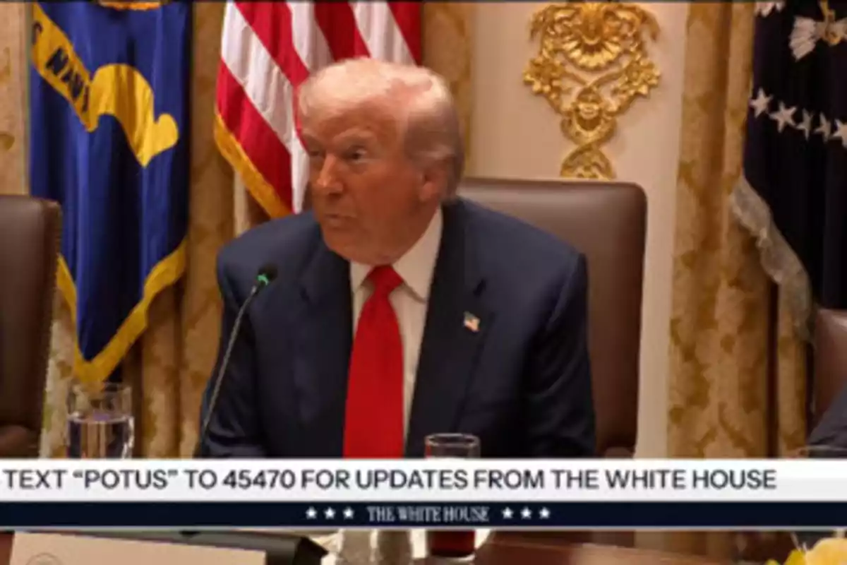 Hombre de cabello rubio con traje azul y corbata roja sentado en una mesa con banderas y decoración oficial de fondo