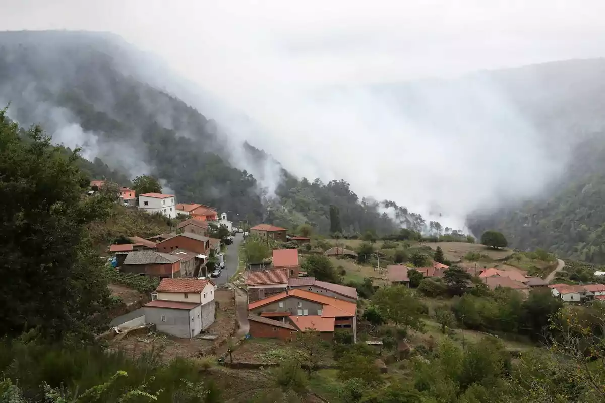 Un pequeño pueblo rodeado de montañas y vegetación con humo cubriendo parte del paisaje debido a un incendio forestal cercano Un pequeño pueblo rodeado de montañas y vegetación con humo cubriendo parte del paisaje debido a un incendio forestal cercano