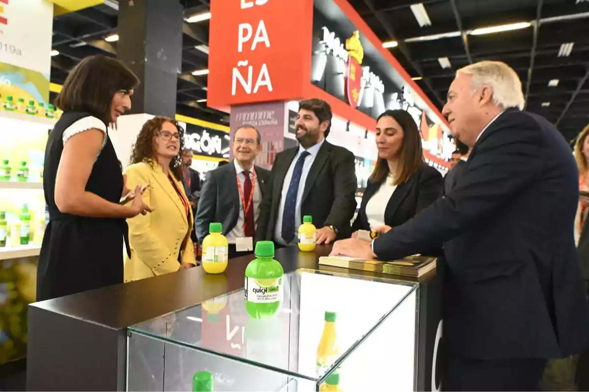 Un grupo de personas vestidas de manera formal conversa alrededor de un mostrador con botellas de jugo en una feria o exposición comercial