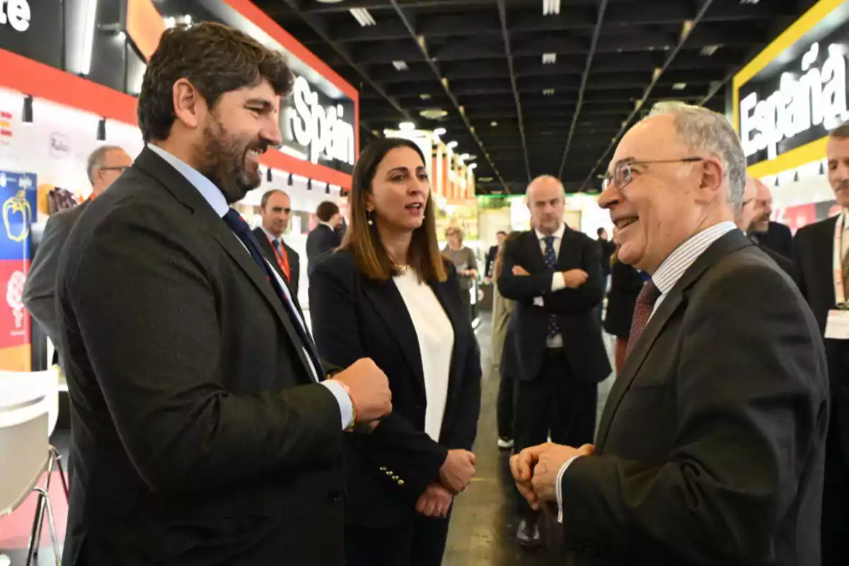 Varias personas vestidas de traje conversan y sonríen en una feria o evento empresarial con letreros de España al fondo