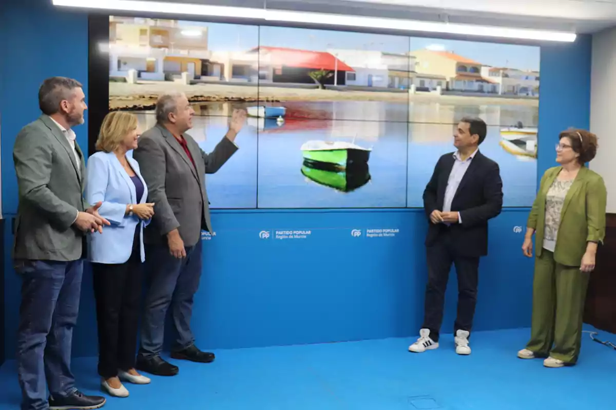 Cinco personas observan una pantalla grande que muestra una imagen de un bote en el agua frente a casas, en una sala con paredes y suelo azules decorados con el logo del Partido Popular Región de Murcia.
