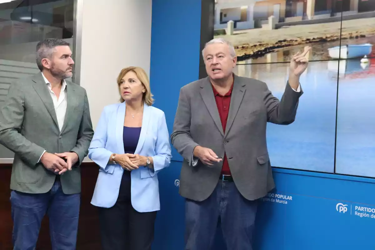 Tres personas de pie con expresión seria, una de ellas señalando una pantalla con una imagen de casas y agua, en un entorno de oficina con fondo azul y logotipos del Partido Popular.
