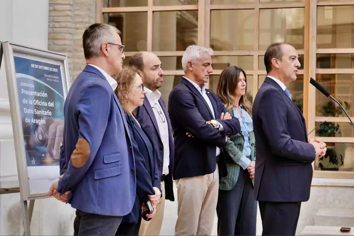 Un grupo de personas vestidas de manera formal está de pie junto a un cartel informativo mientras un hombre habla frente a un micrófono en un evento oficial en un edificio con ventanas grandes.