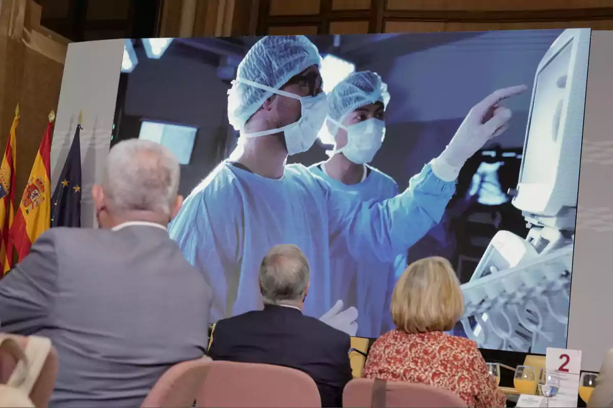 Personas sentadas observan una pantalla grande que muestra a dos médicos con mascarilla y gorro quirúrgico trabajando en un hospital Personas sentadas observan una pantalla grande que muestra a dos médicos con mascarilla y gorro quirúrgico trabajando en un hospital