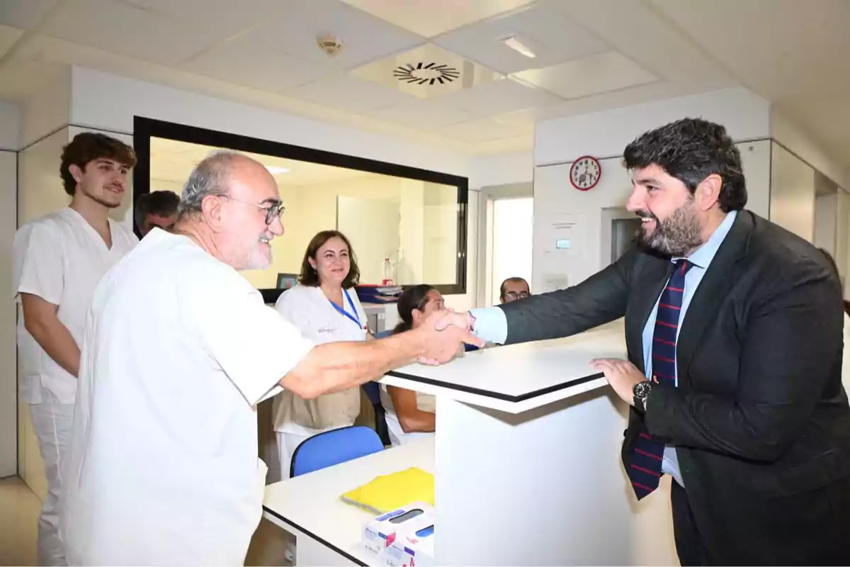 Un hombre con bata blanca y un hombre con traje se dan la mano en una recepción de hospital mientras otras personas observan sonrientes. Un hombre con bata blanca y un hombre con traje se dan la mano en una recepción de hospital mientras otras personas observan sonrientes.