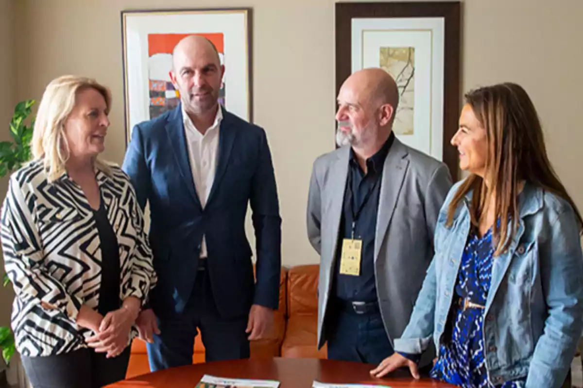 Cuatro personas de pie conversando en una oficina, dos hombres y dos mujeres, con cuadros en la pared y documentos sobre la mesa. Cuatro personas de pie conversando en una oficina, dos hombres y dos mujeres, con cuadros en la pared y documentos sobre la mesa.