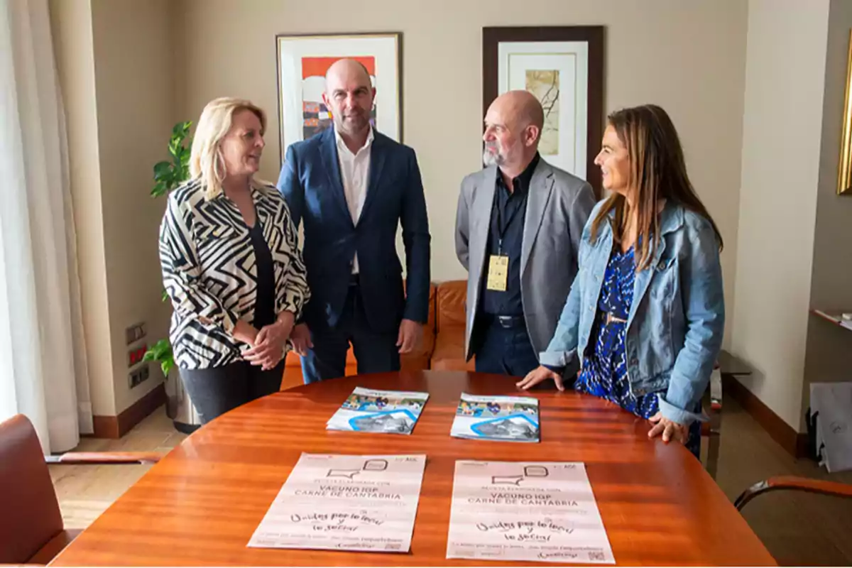 Cuatro personas de pie alrededor de una mesa de madera en una oficina, conversando y sonriendo, con folletos y carteles sobre la mesa y cuadros en la pared de fondo.