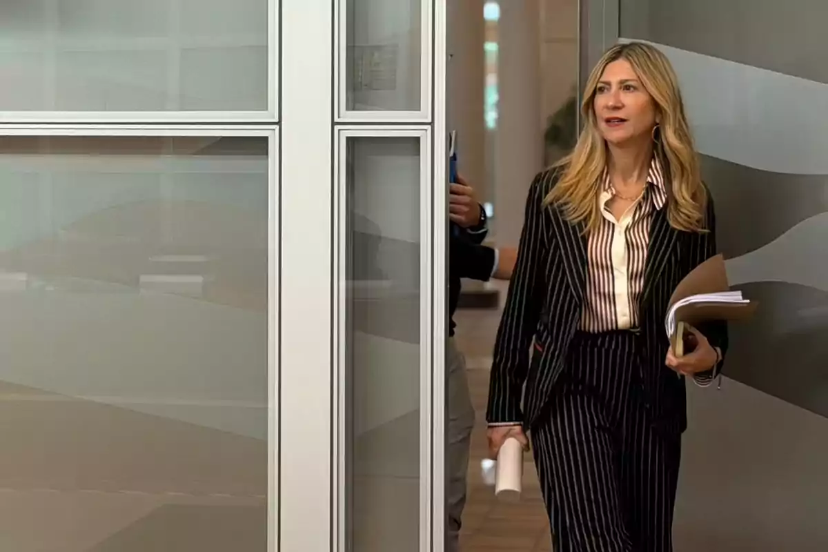 Mujer rubia con traje de rayas entra a una oficina sosteniendo documentos y una carpeta