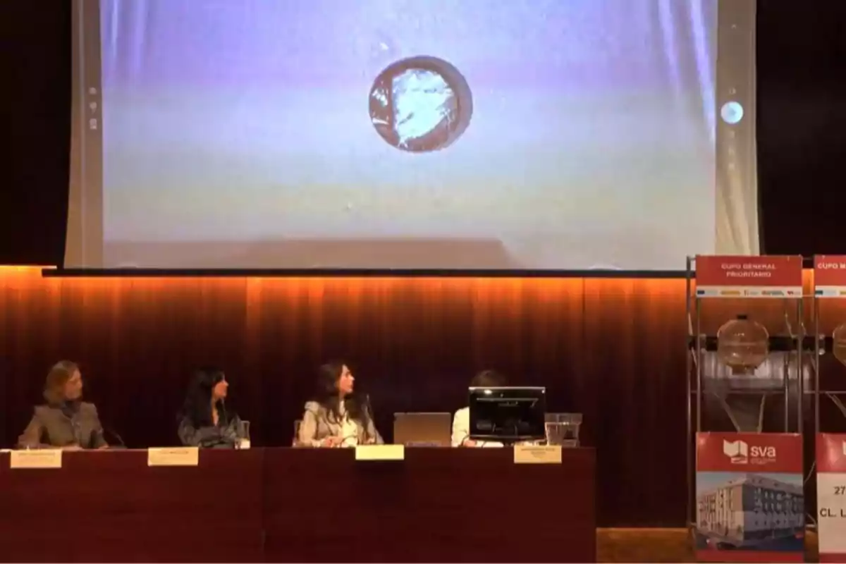 Cuatro personas sentadas en una mesa de conferencias observan una pantalla grande donde se proyecta una imagen circular, con urnas transparentes a la derecha del escenario.